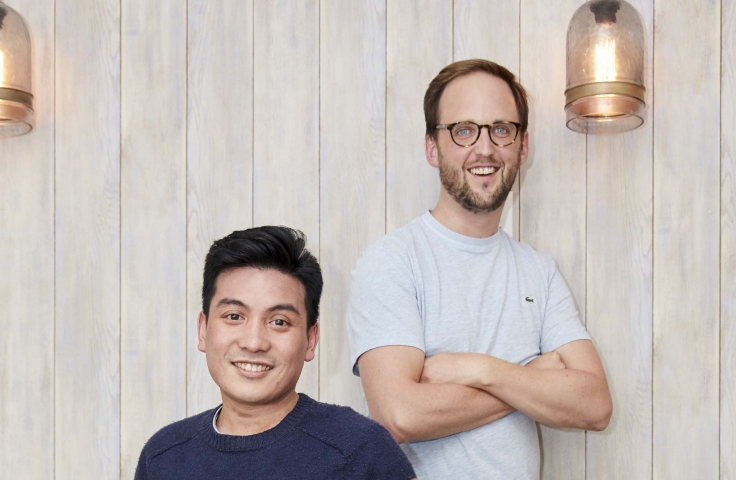 The founders of Forage, Pasha Rayan and Tom Brunskill, both male. One sits on the edge of a sofa and the other stands cross armed, in front of a wooden wall. 